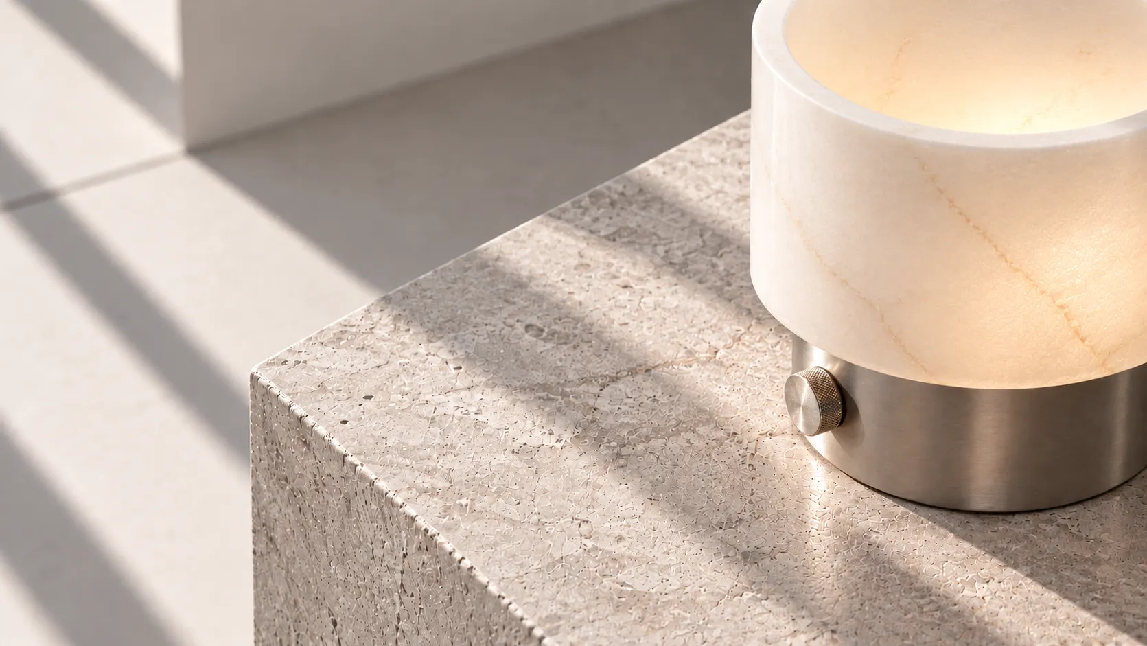 Raking sunlight across a stone plinth, on which a veined alabaster cylinder glows above a brushed metal base.